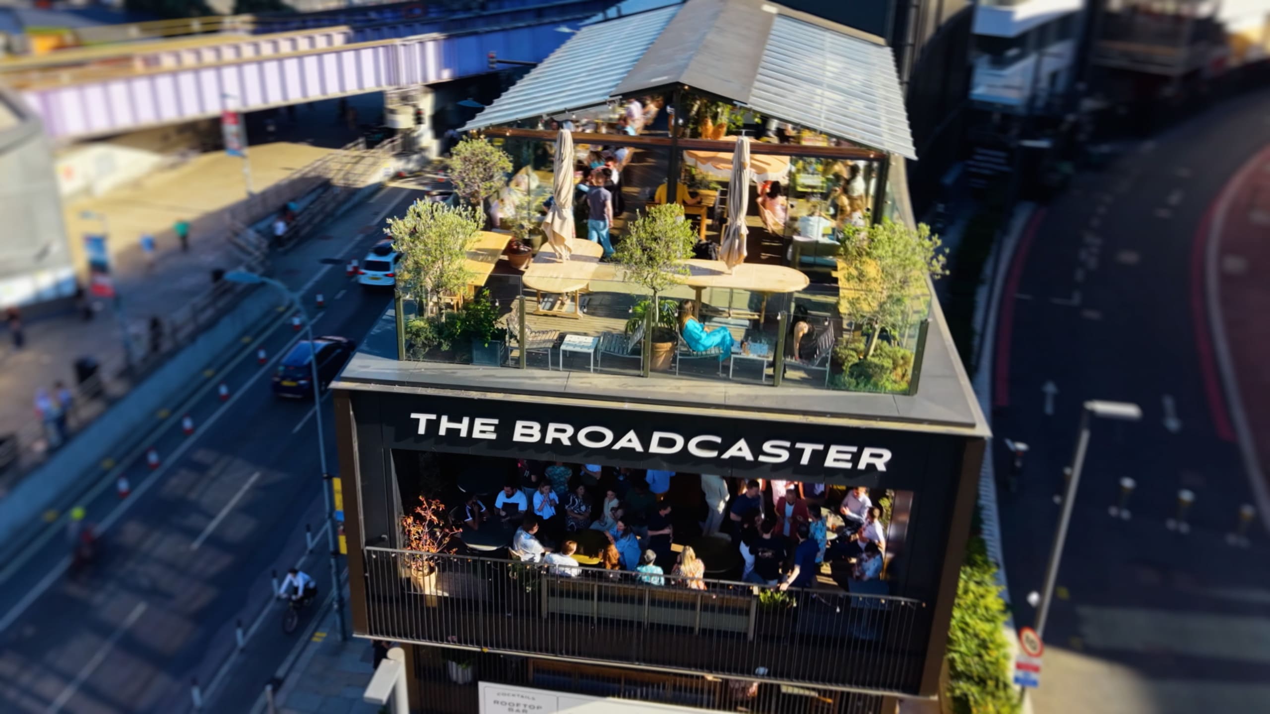 Aerial view of "The Broadcaster" building with a busy rooftop terrace filled with people and greenery, next to a city street and rail bridge.