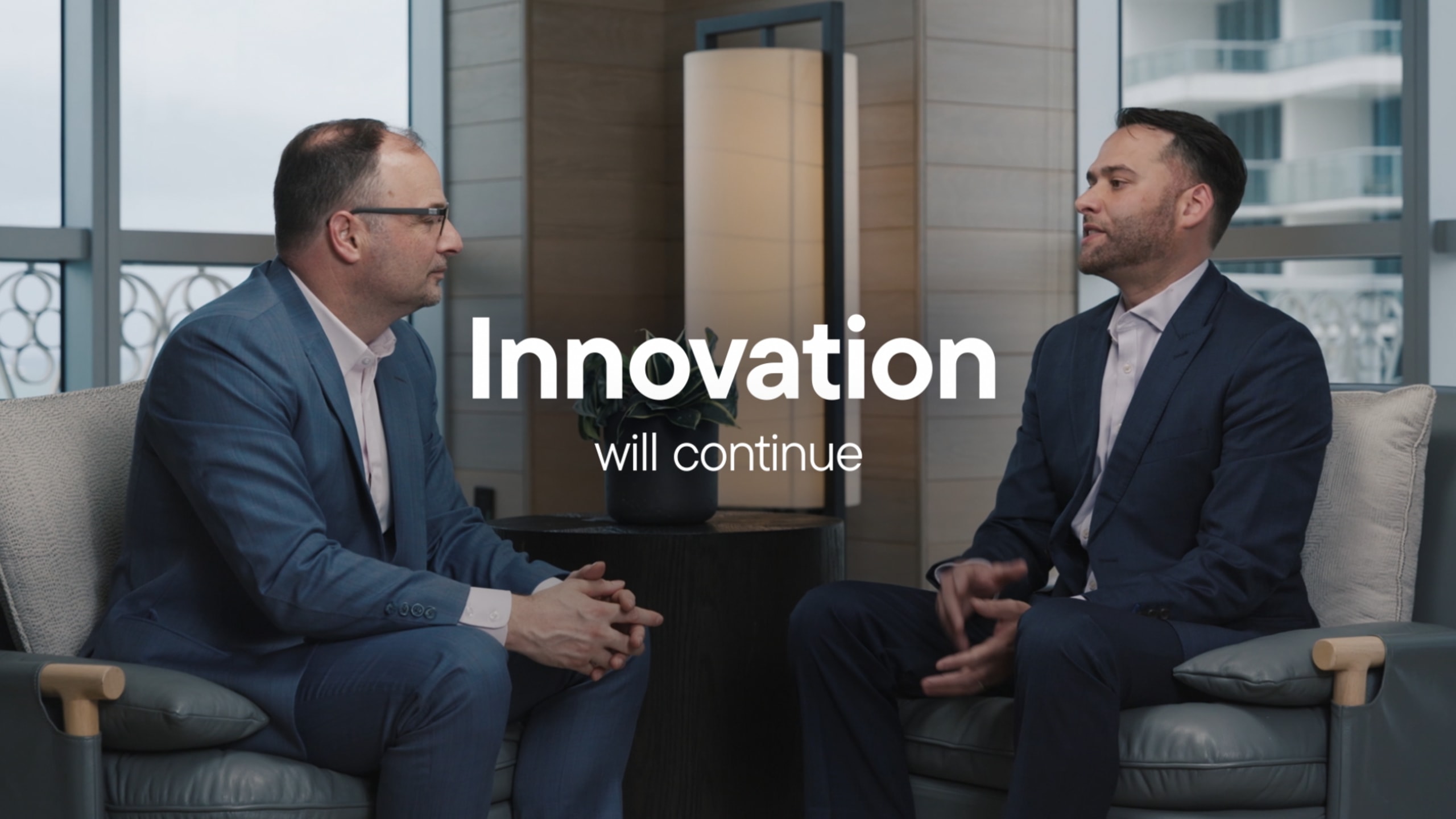 Two men in suits sit facing each other in an office setting. Text overlay reads "Innovation will continue."