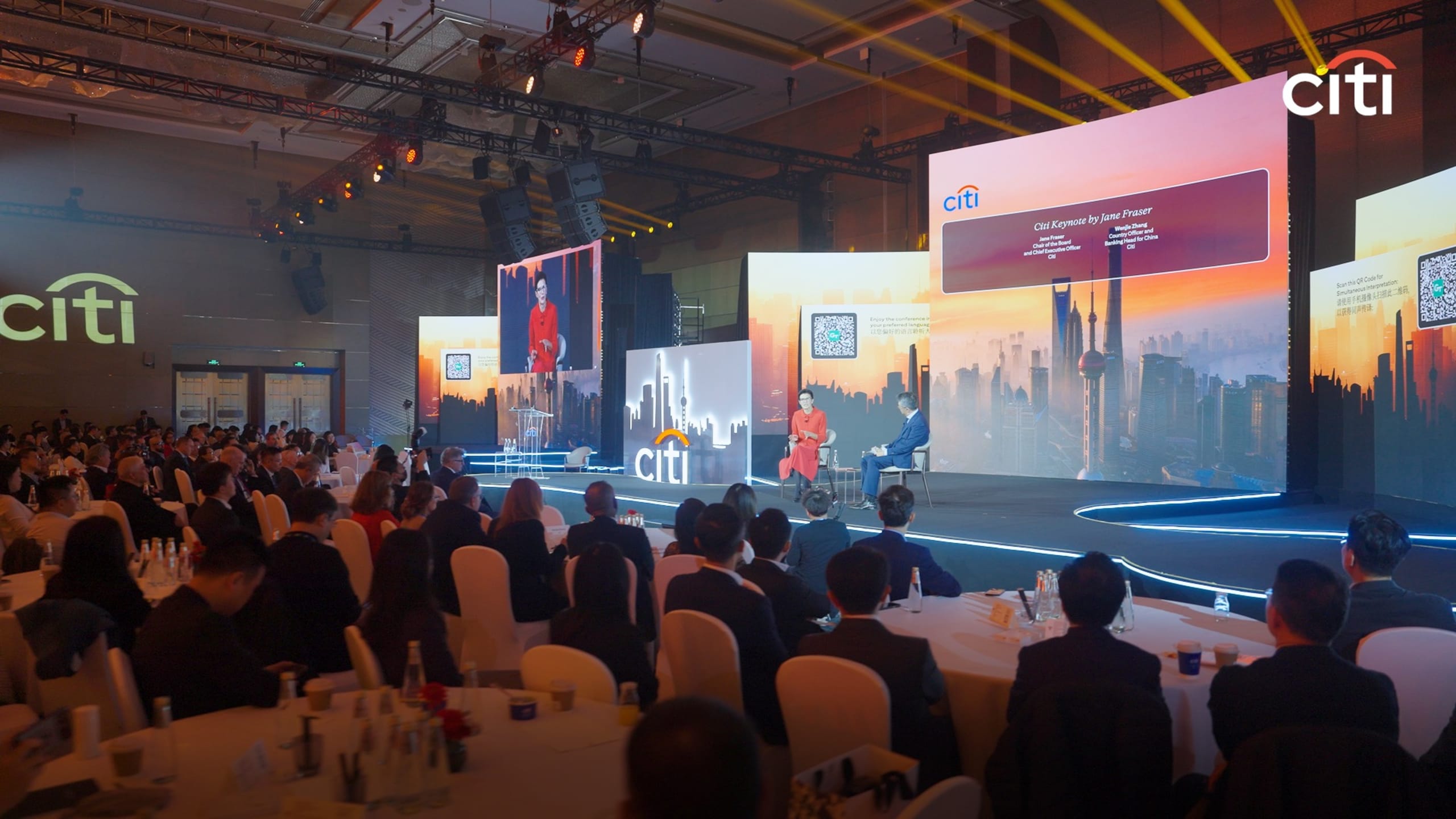 Audience at a Citi event with a speaker on stage, large screens displaying cityscape visuals, and branded logos.