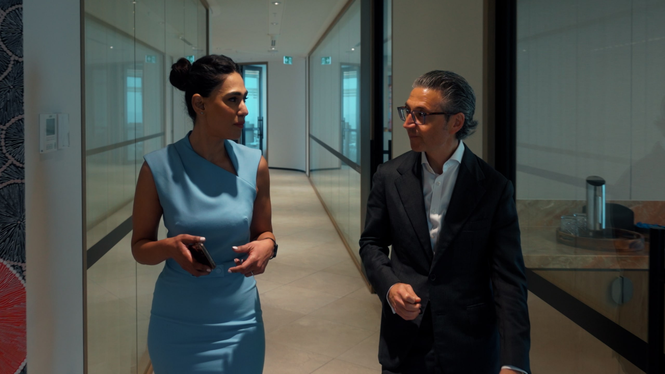A woman in a blue dress and a man in a black suit walk and talk in a modern office hallway, holding a phone and engaged in conversation.
