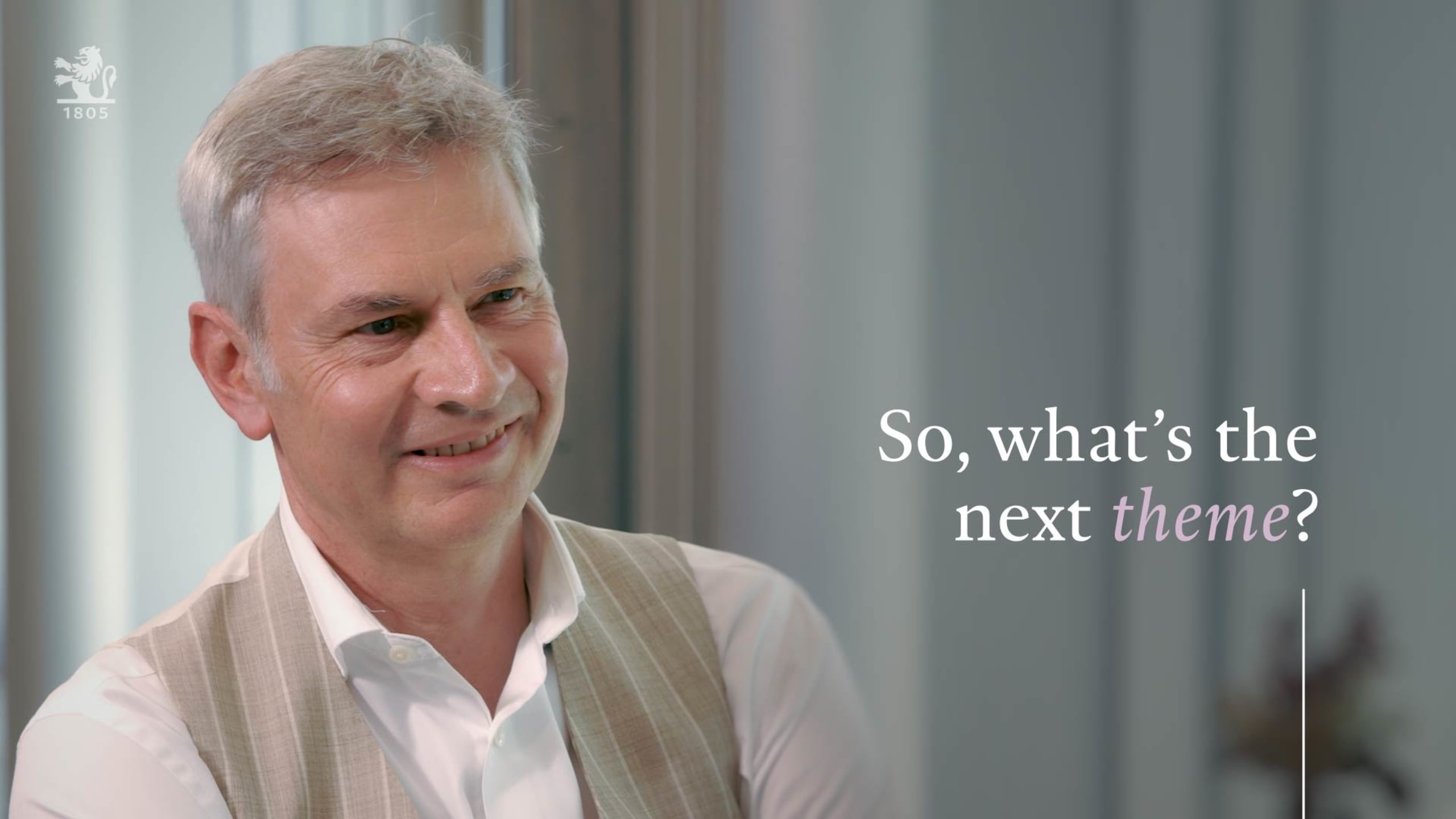 Smiling man in a light vest and white shirt with text overlay: "So, what's the next theme?" in a softly lit office setting.