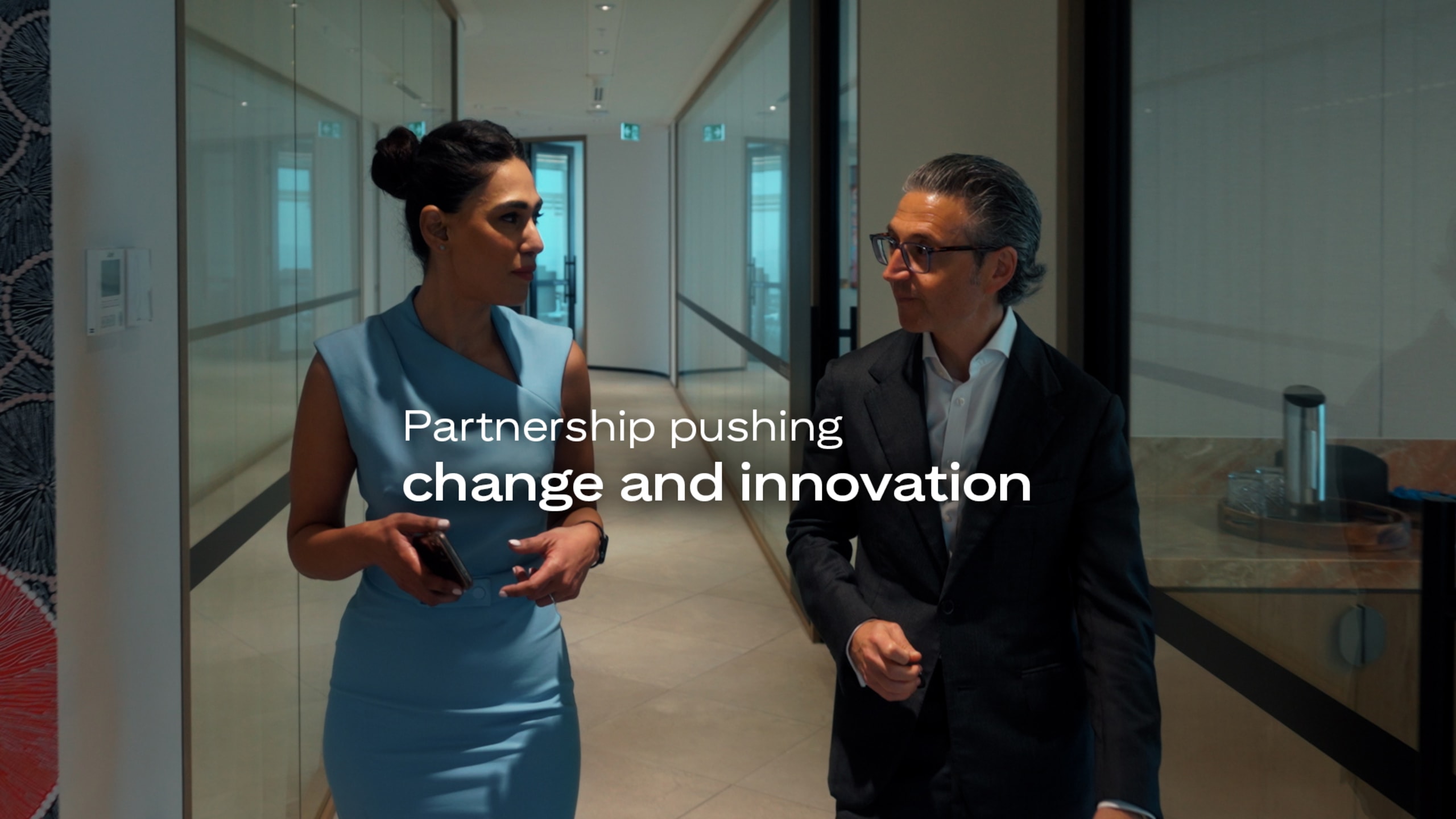 Two professionals walk in an office corridor, engaged in discussion, with "Partnership pushing change and innovation" text overlay.