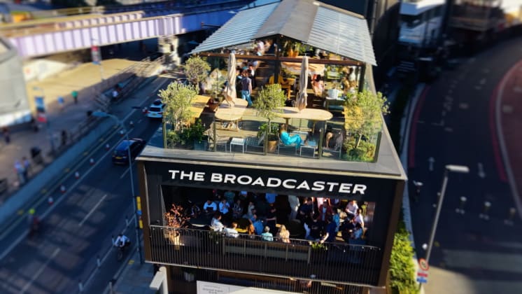 Aerial view of "The Broadcaster" building with a busy rooftop terrace filled with people and greenery, next to a city street and rail bridge.