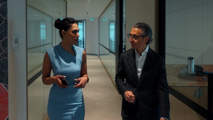 A woman in a blue dress and a man in a black suit walk and talk in a modern office hallway, holding a phone and engaged in conversation.