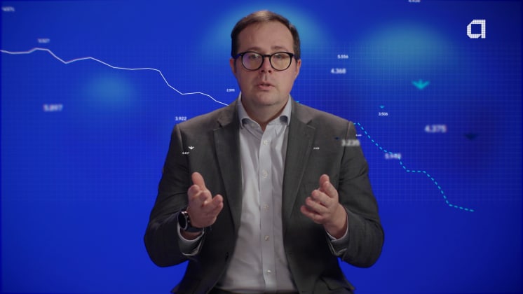 A man in a suit and glasses gestures while sitting. Behind him, a blue background displays a declining line graph with numerical data.
