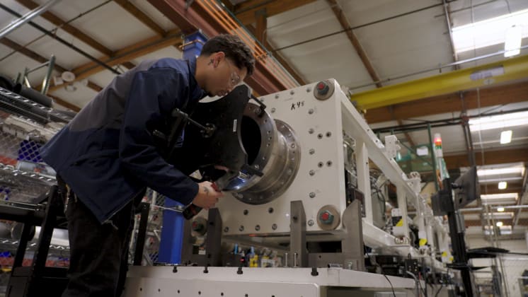 A person in safety glasses operates a large industrial machine in a factory setting, surrounded by metal structures and equipment.